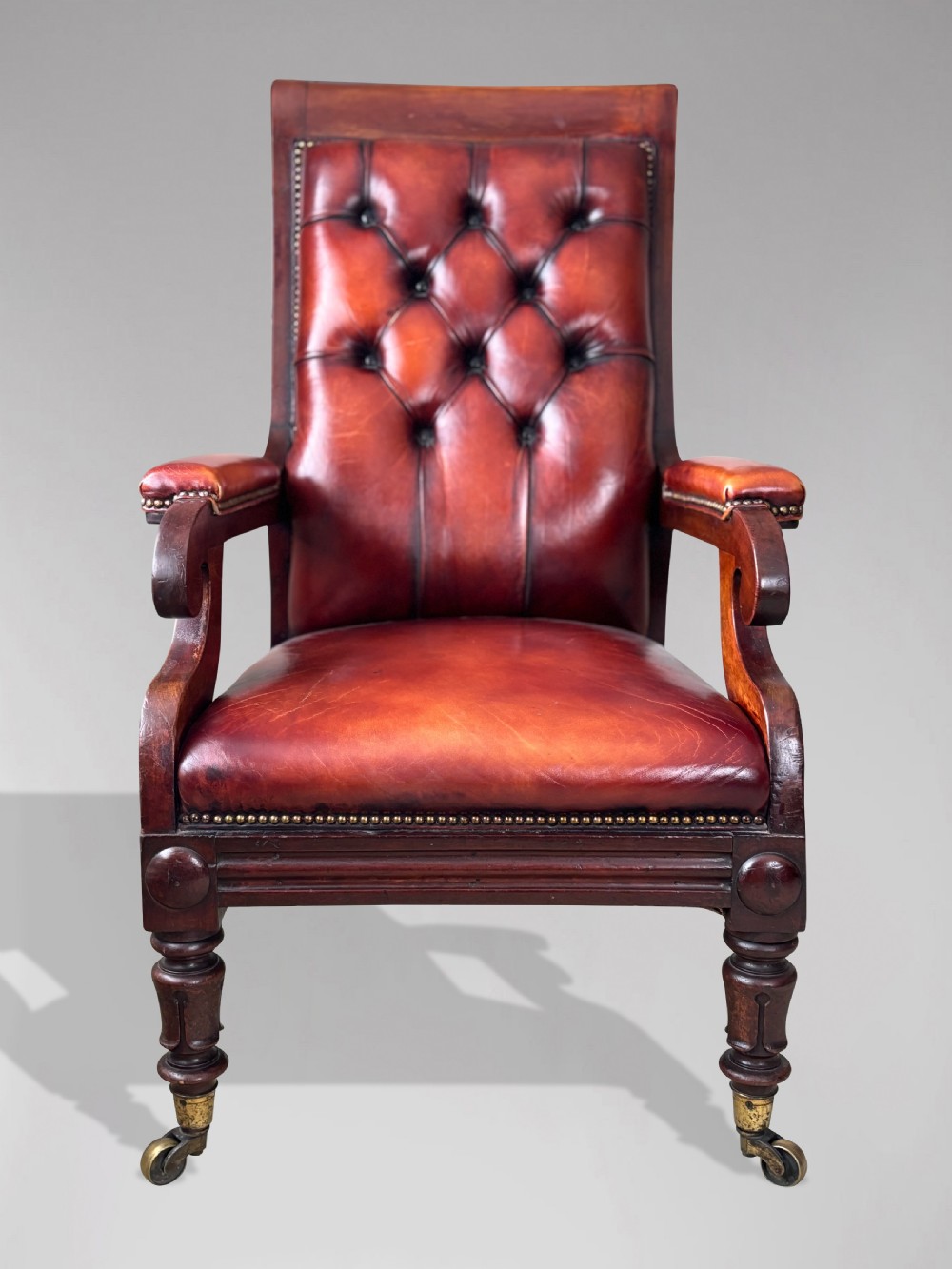 A 19th-century William IV mahogany library armchair featuring deep-buttoned red-brown leather upholstery, scrolled arms, brass stud trim, and turned front legs on brass castors. View IV