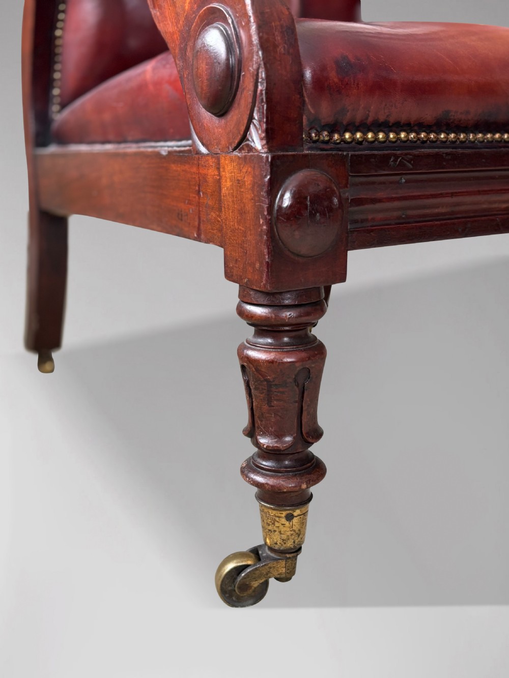 A 19th-century William IV mahogany library armchair featuring deep-buttoned red-brown leather upholstery, scrolled arms, brass stud trim, and turned front legs on brass castors. View V