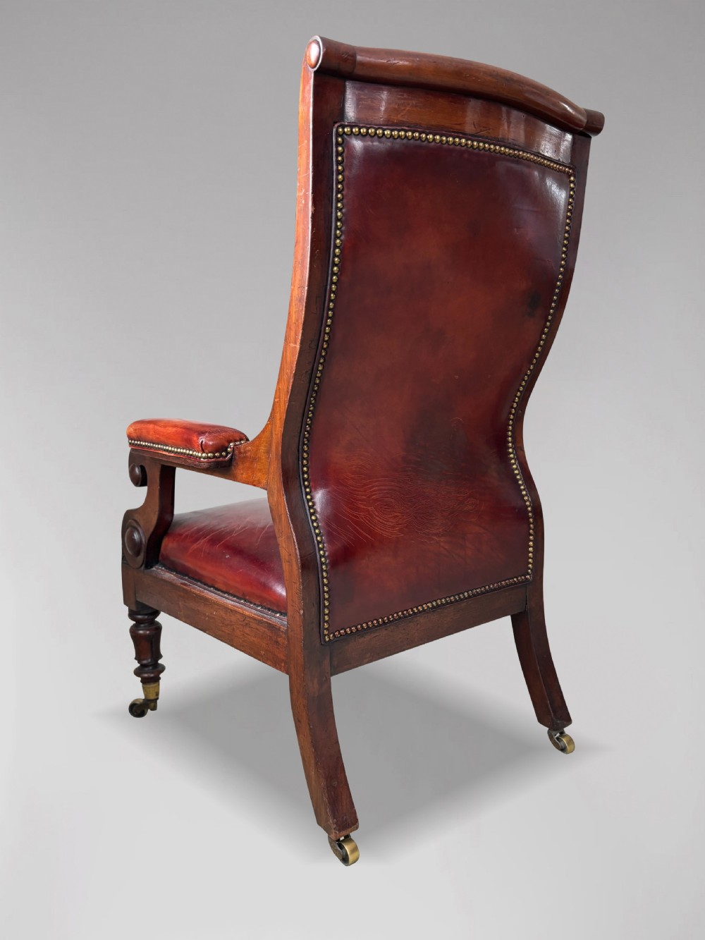 A 19th-century William IV mahogany library armchair featuring deep-buttoned red-brown leather upholstery, scrolled arms, brass stud trim, and turned front legs on brass castors. View VI