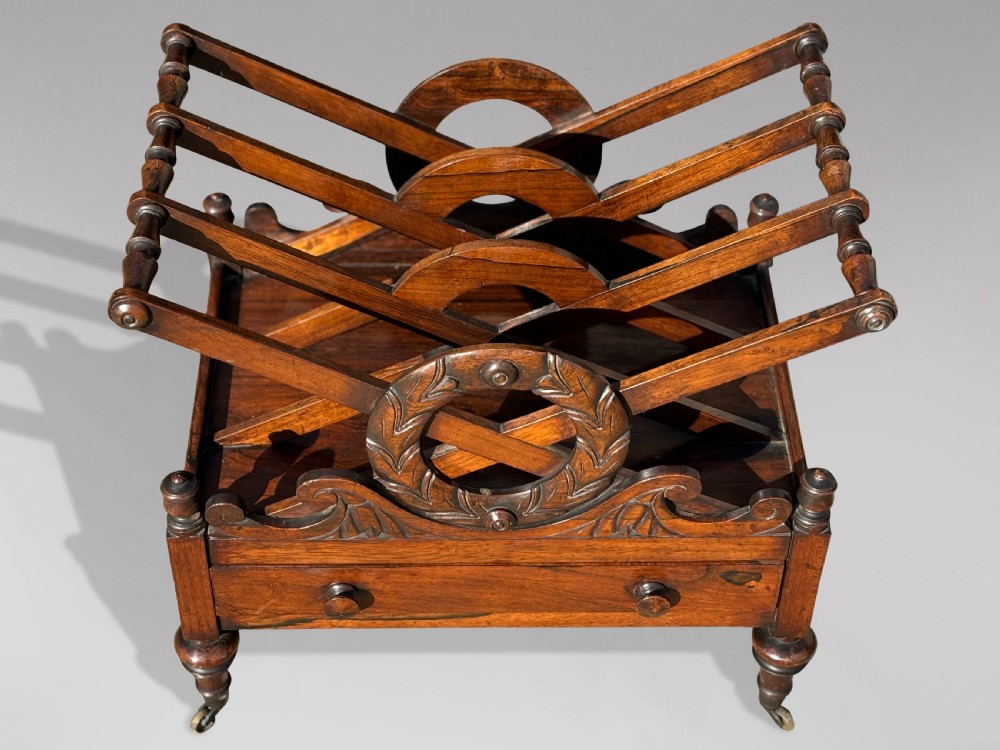 A 19th-century Regency rosewood Canterbury featuring slatted music dividers, carved laurel wreath side handles, a single base drawer, and turned legs with brass castors. View II
