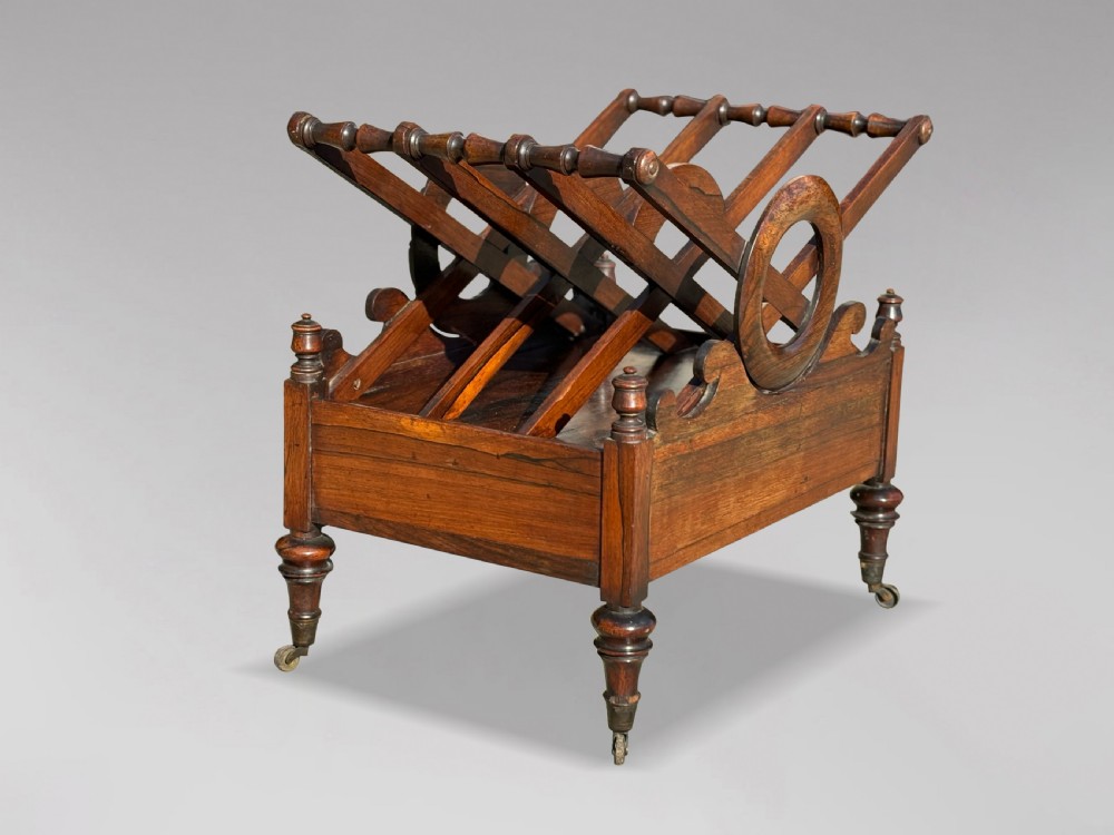 A 19th-century Regency rosewood Canterbury featuring slatted music dividers, carved laurel wreath side handles, a single base drawer, and turned legs with brass castors. View V