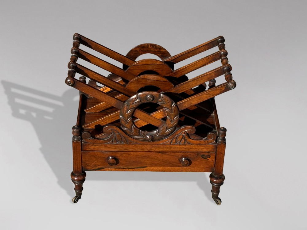 A 19th-century Regency rosewood Canterbury featuring slatted music dividers, carved laurel wreath side handles, a single base drawer, and turned legs with brass castors. View VI