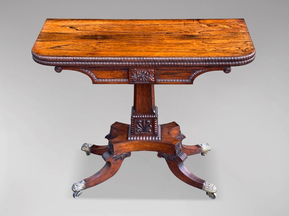 A 19th-century George IV period rosewood fold-over card table featuring a figured veneer top, carved pedestal base, and four splayed legs with brass paw castors.