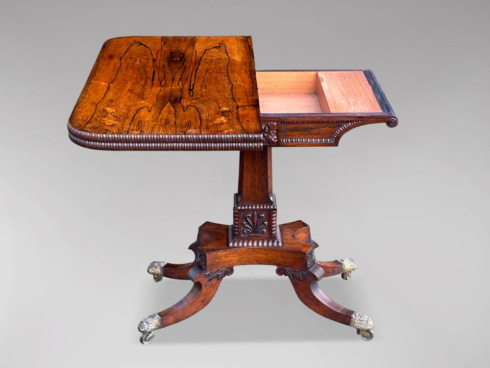 A 19th-century George IV period rosewood fold-over card table featuring a figured veneer top, carved pedestal base, and four splayed legs with brass paw castors. View II