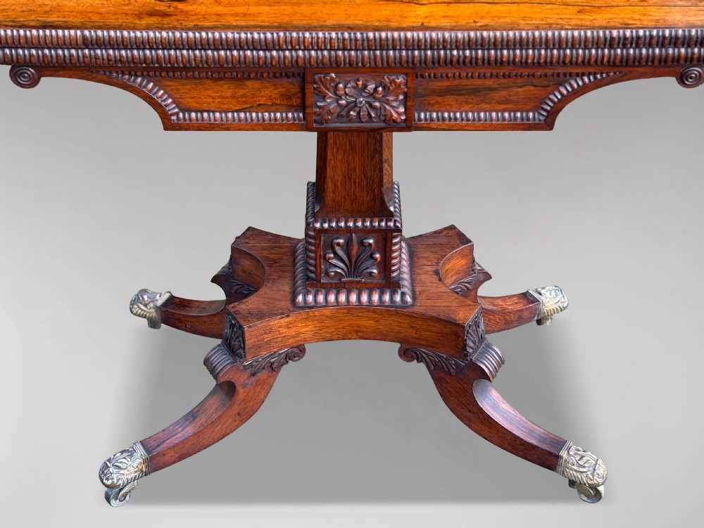 A 19th-century George IV period rosewood fold-over card table featuring a figured veneer top, carved pedestal base, and four splayed legs with brass paw castors. View IV