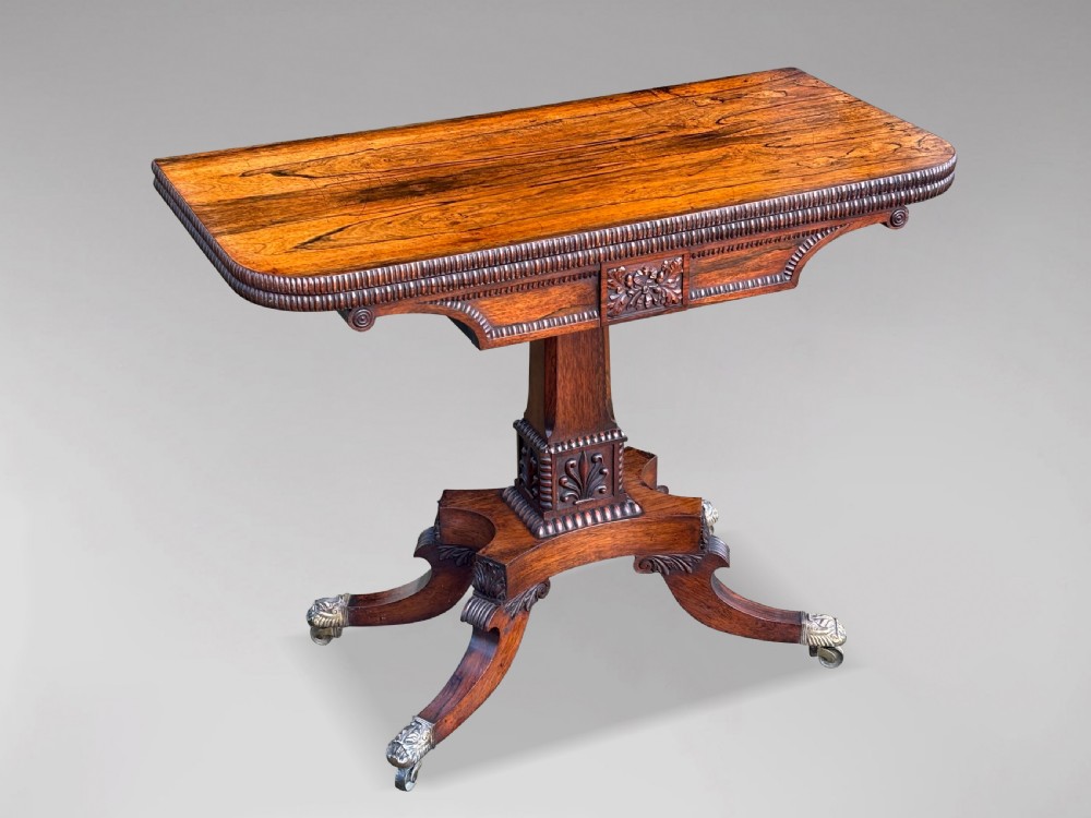 A 19th-century George IV period rosewood fold-over card table featuring a figured veneer top, carved pedestal base, and four splayed legs with brass paw castors. View X