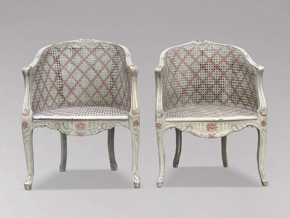 A pair of 20th-century French Louis XV style armchairs with off-white painted frames, pink floral carvings, cabriole legs, and woven cane backs adorned with a painted red diamond pattern.