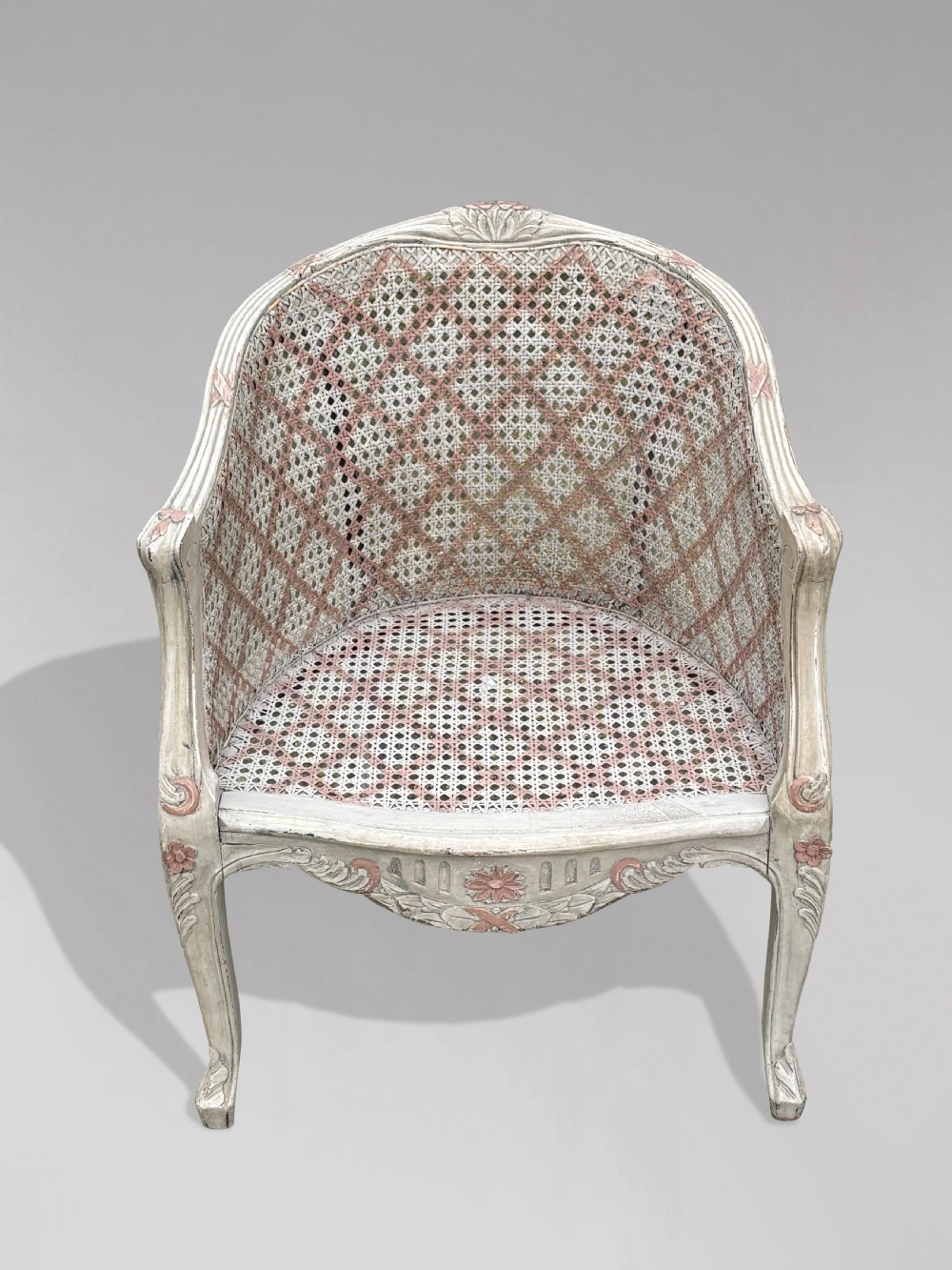 A pair of 20th-century French Louis XV style armchairs with off-white painted frames, pink floral carvings, cabriole legs, and woven cane backs adorned with a painted red diamond pattern. View V