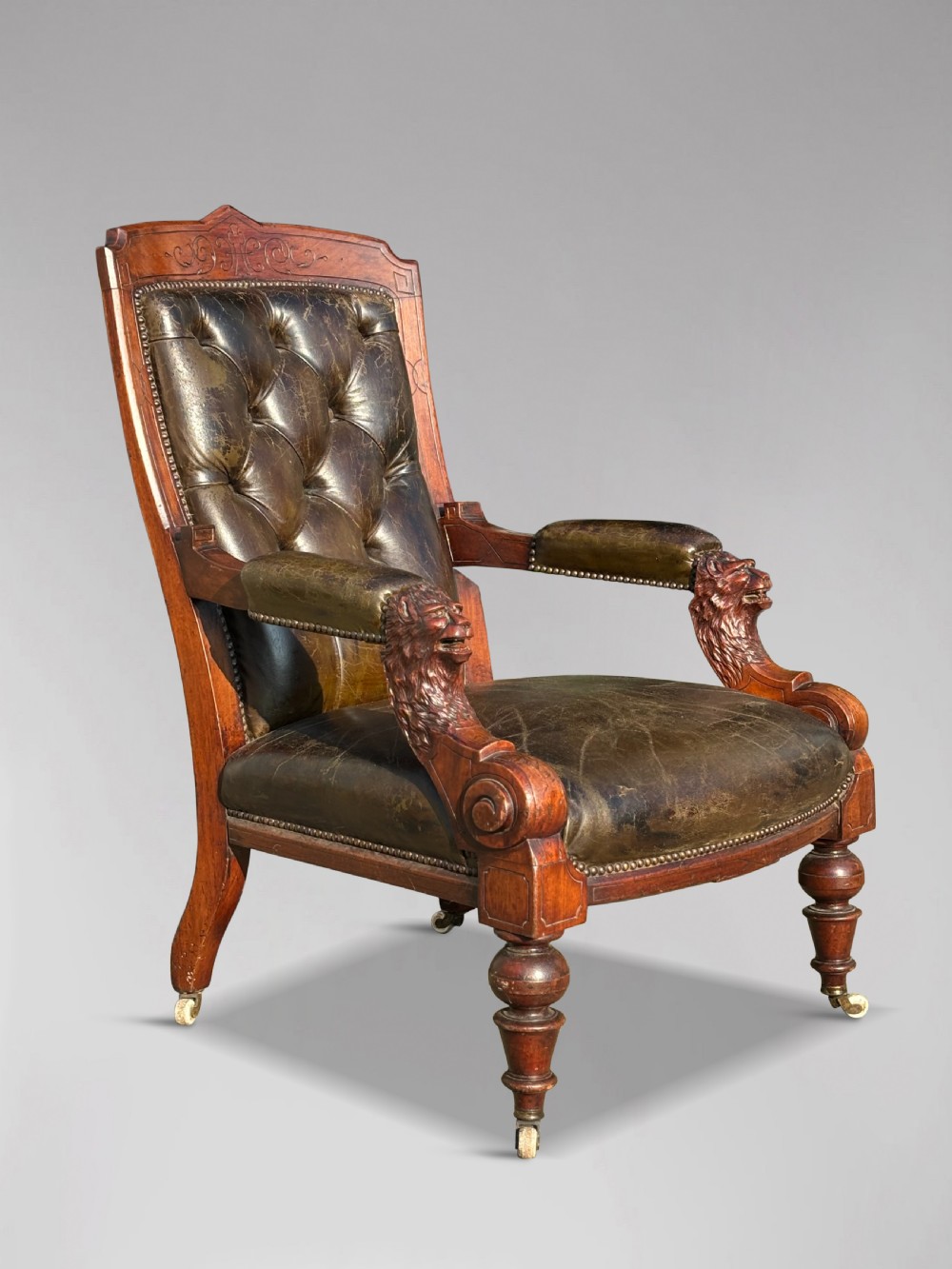 A nineteenth-century carved walnut library armchair with button-tufted dark green leather upholstery, carved lion-head armrests, and turned legs with castors.