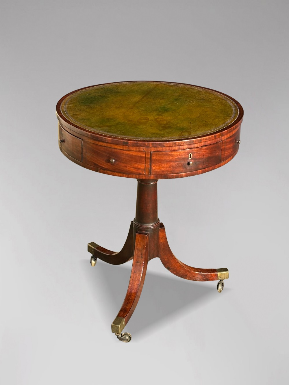 An 18th-century George III mahogany drum table with a circular green leather top, frieze drawers, turned pedestal, and splayed legs with brass castors.