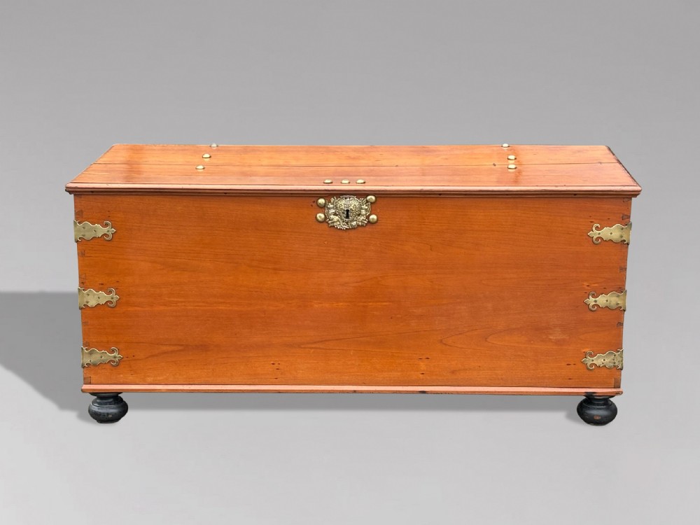 An 18th-century warm brown walnut colonial chest featuring brass corner brackets, a central decorative brass escutcheon, lid studs, and dark turned bun feet.