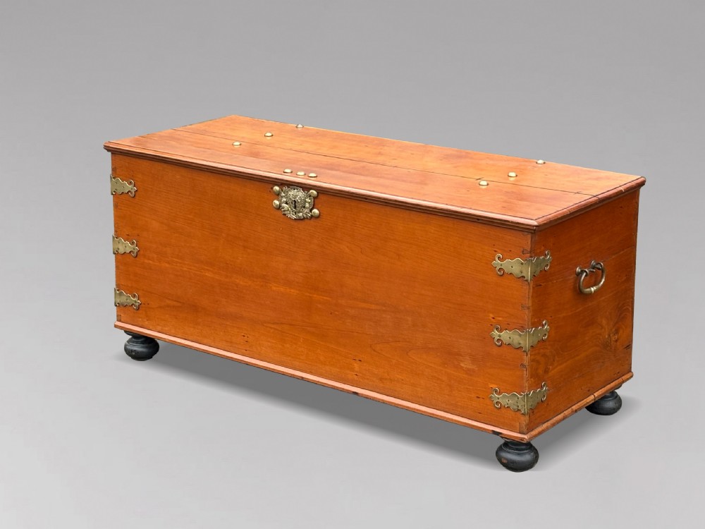 An 18th-century warm brown walnut colonial chest featuring brass corner brackets, a central decorative brass escutcheon, lid studs, and dark turned bun feet. View III