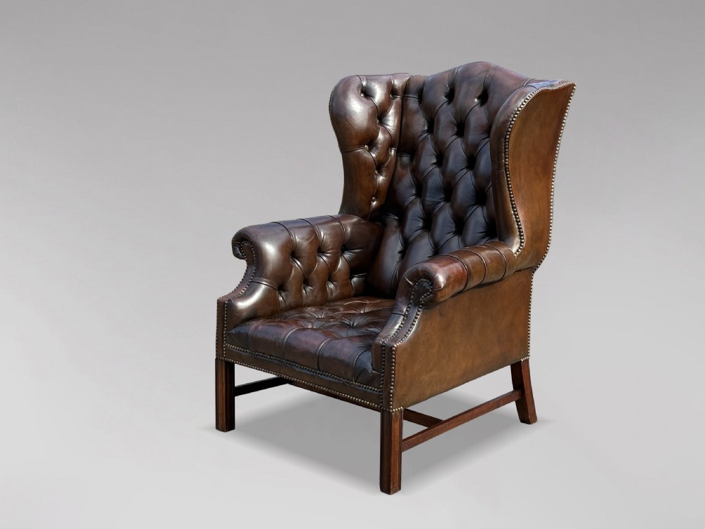 A dark brown leather wingback armchair featuring deep button tufting, brass studding along the front edges, and square timber legs with stretchers.