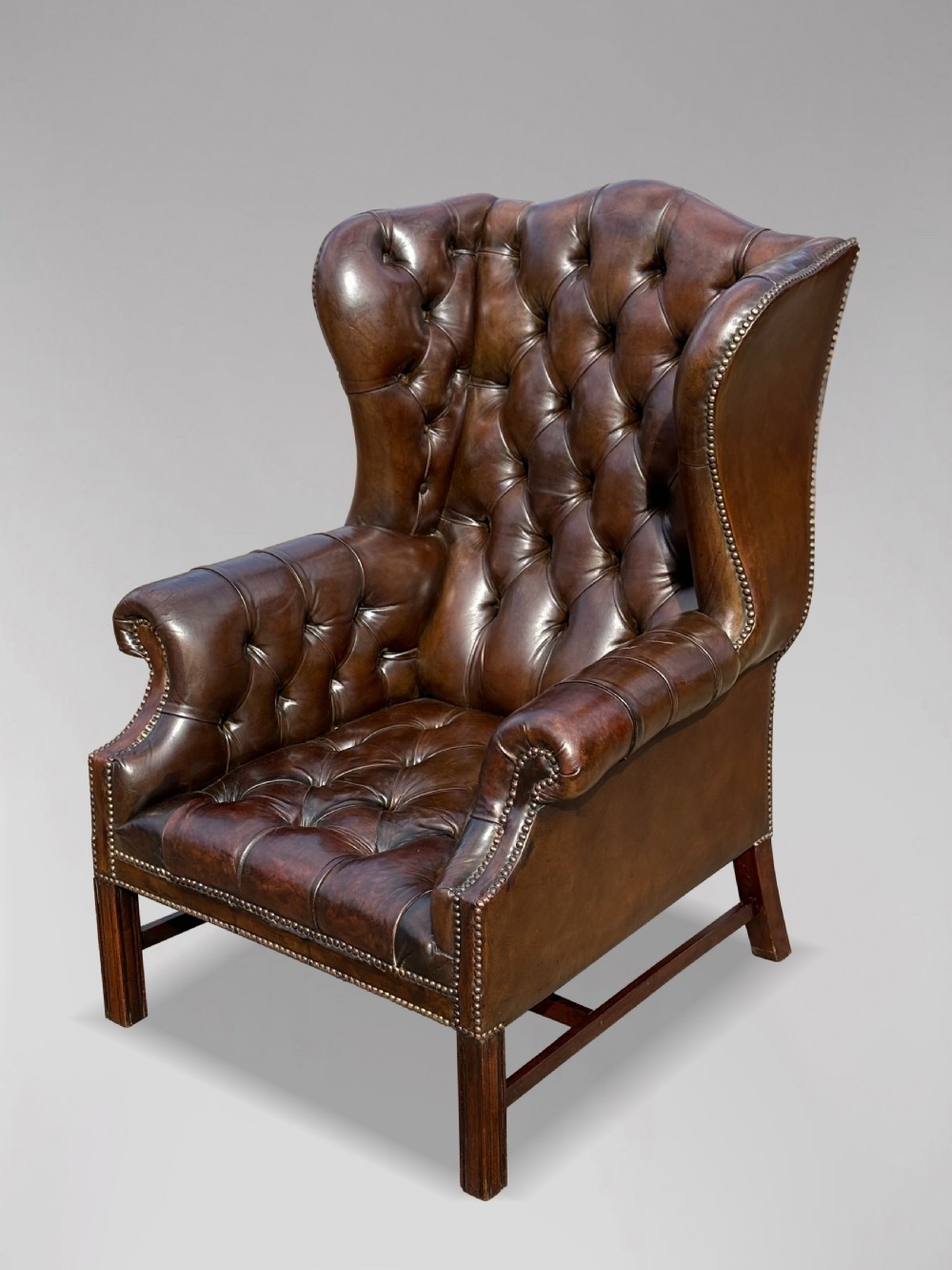 A dark brown leather wingback armchair featuring deep button tufting, brass studding along the front edges, and square timber legs with stretchers. View II