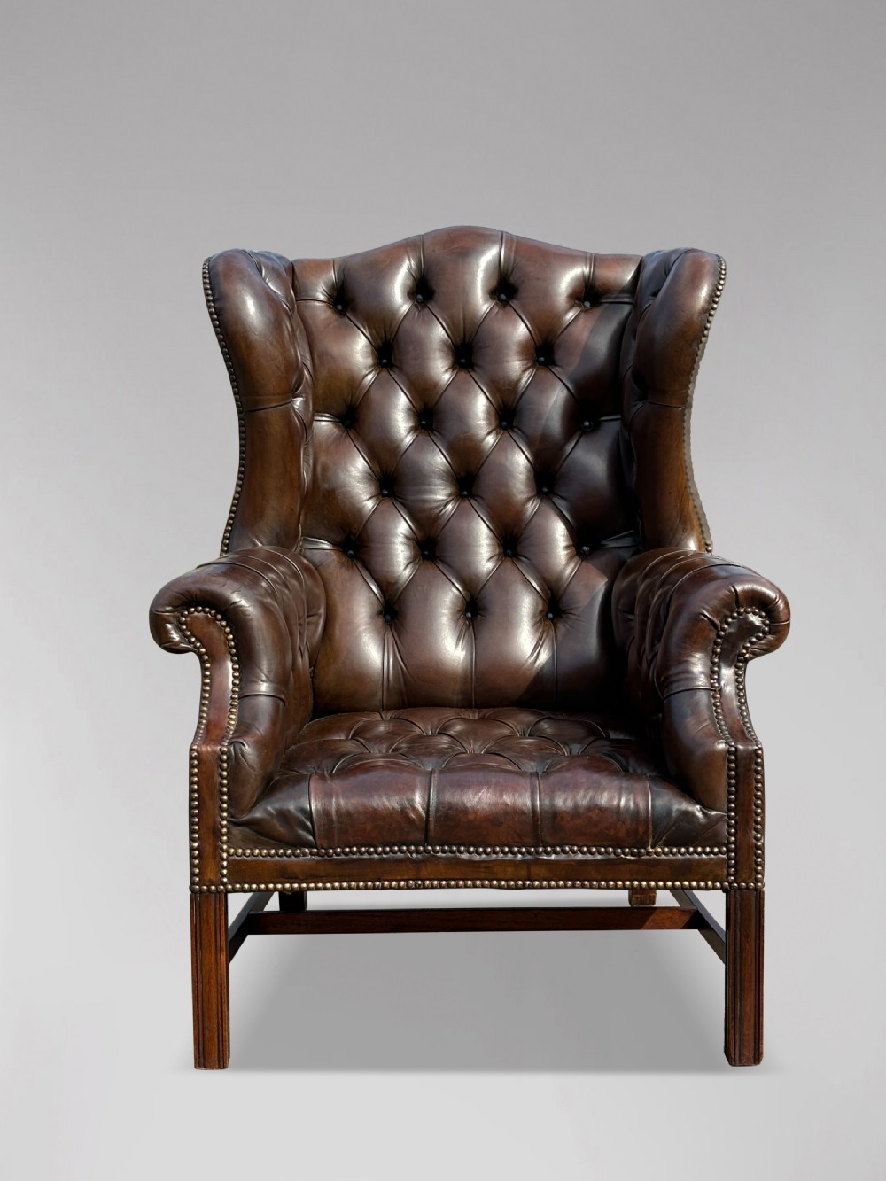 A dark brown leather wingback armchair featuring deep button tufting, brass studding along the front edges, and square timber legs with stretchers. View III