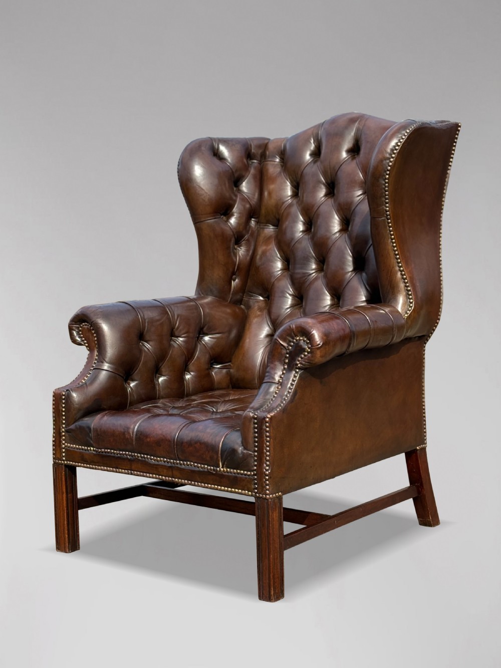 A dark brown leather wingback armchair featuring deep button tufting, brass studding along the front edges, and square timber legs with stretchers. View IV