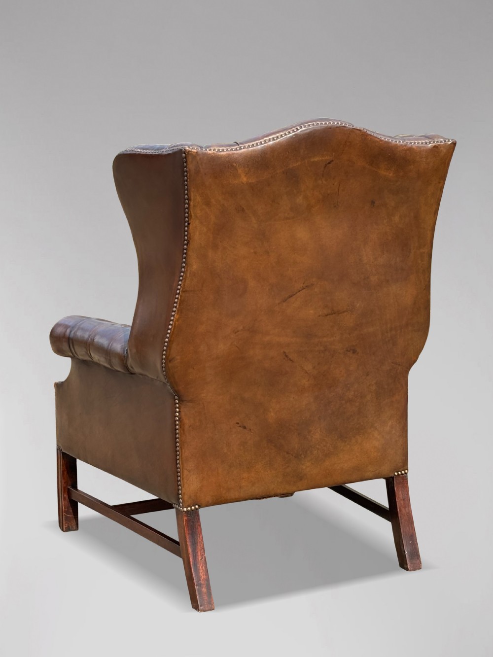 A dark brown leather wingback armchair featuring deep button tufting, brass studding along the front edges, and square timber legs with stretchers. View V