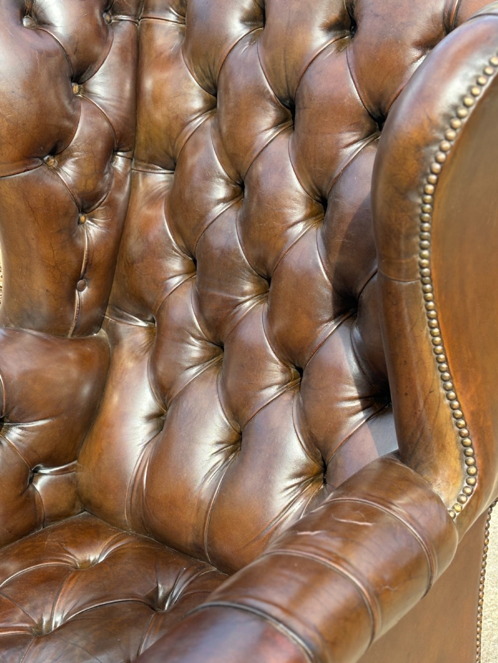 A dark brown leather wingback armchair featuring deep button tufting, brass studding along the front edges, and square timber legs with stretchers. View VI