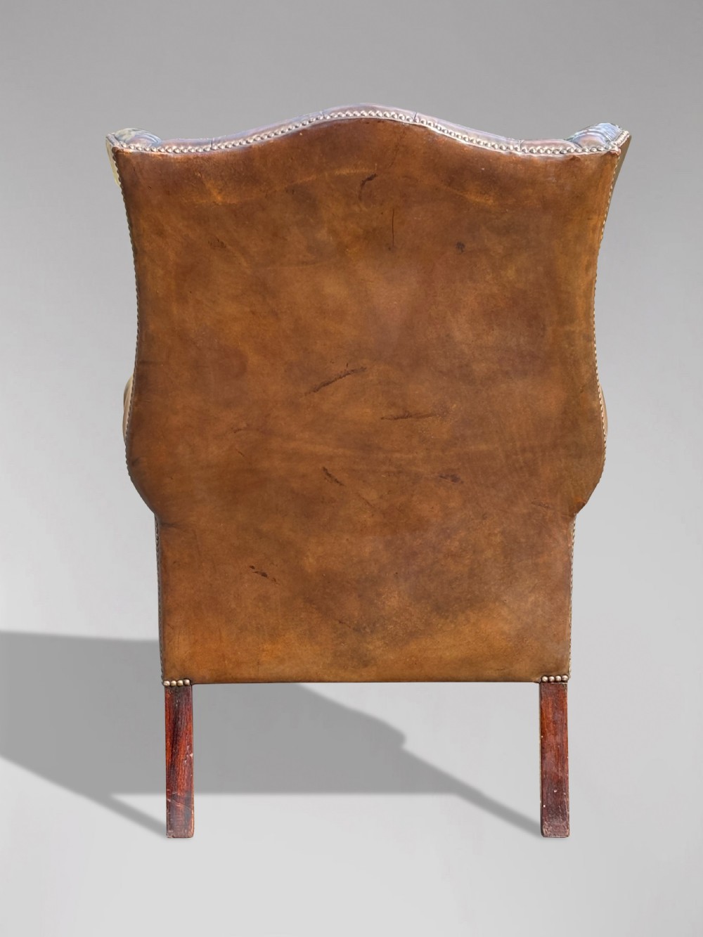 A dark brown leather wingback armchair featuring deep button tufting, brass studding along the front edges, and square timber legs with stretchers. View VII