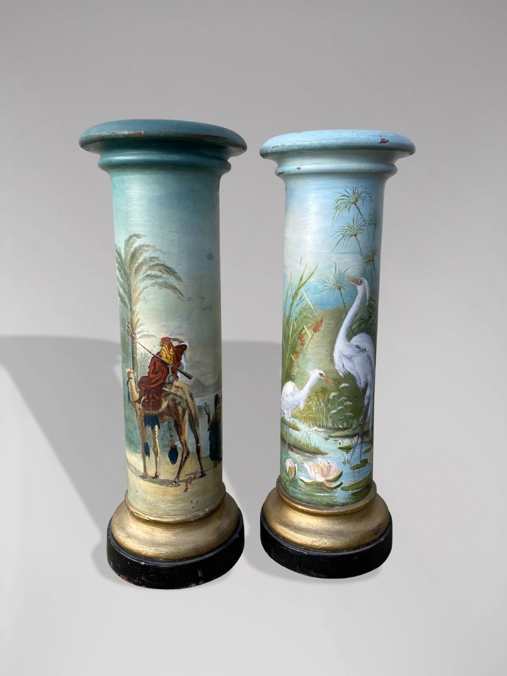 A pair of 19th-century French hand-painted cylindrical wooden columns featuring narrative desert and riverbank scenes above turned gilded bases.