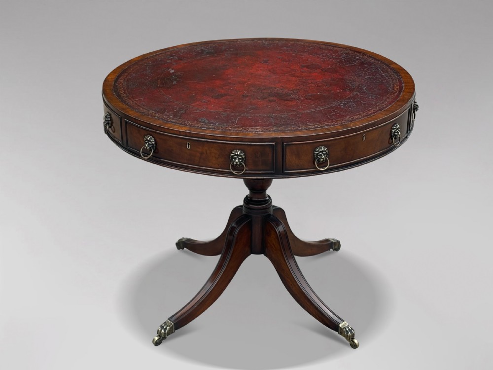 A 19th-century circular mahogany drum table featuring a red leather inlaid top, brass lion mask handles on perimeter drawers, and a pedestal base on splayed legs with brass paw castors.