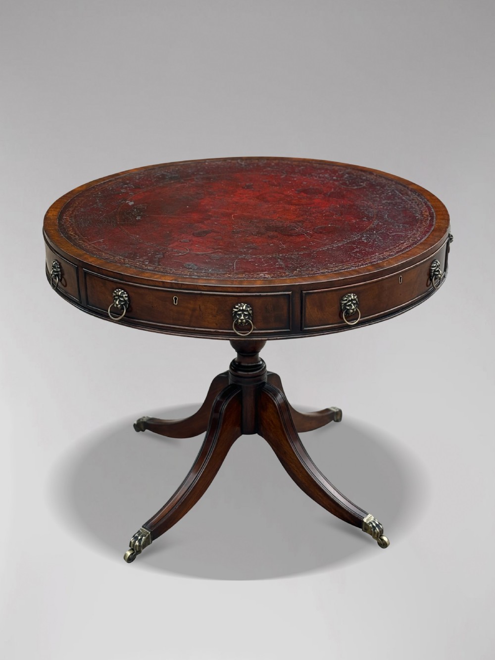 A 19th-century circular mahogany drum table featuring a red leather inlaid top, brass lion mask handles on perimeter drawers, and a pedestal base on splayed legs with brass paw castors. View II