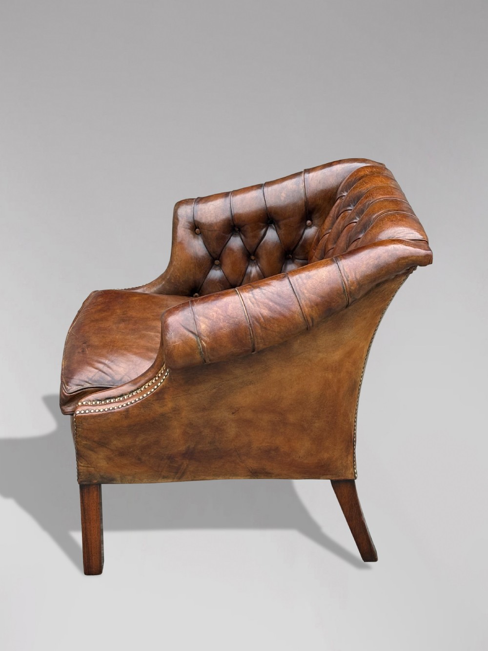 A 19th-century brown leather club armchair featuring deep button tufting, rolled arms with brass studding, and tapered timber legs. View V