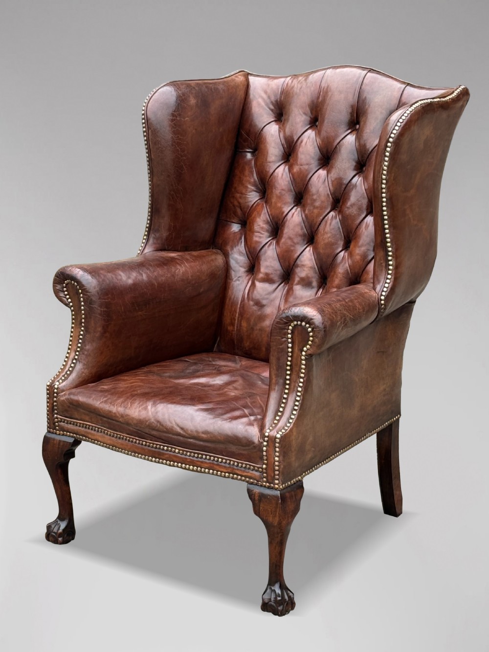 A 19th-century deep brown leather wingback armchair featuring a button-tufted backrest, brass stud trim, and hand-carved mahogany cabriole legs with ball-and-claw feet.
