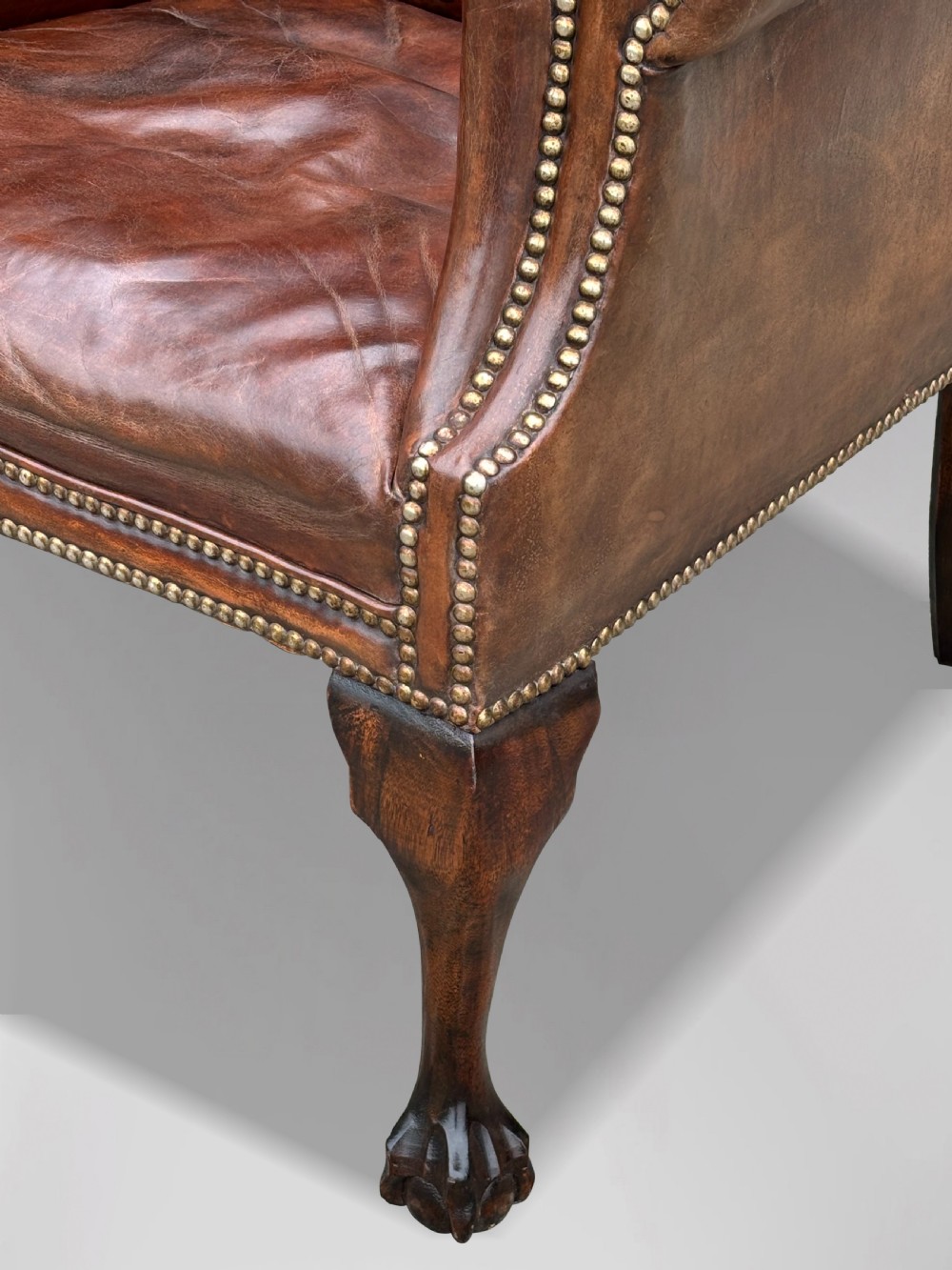 A 19th-century deep brown leather wingback armchair featuring a button-tufted backrest, brass stud trim, and hand-carved mahogany cabriole legs with ball-and-claw feet. View VI