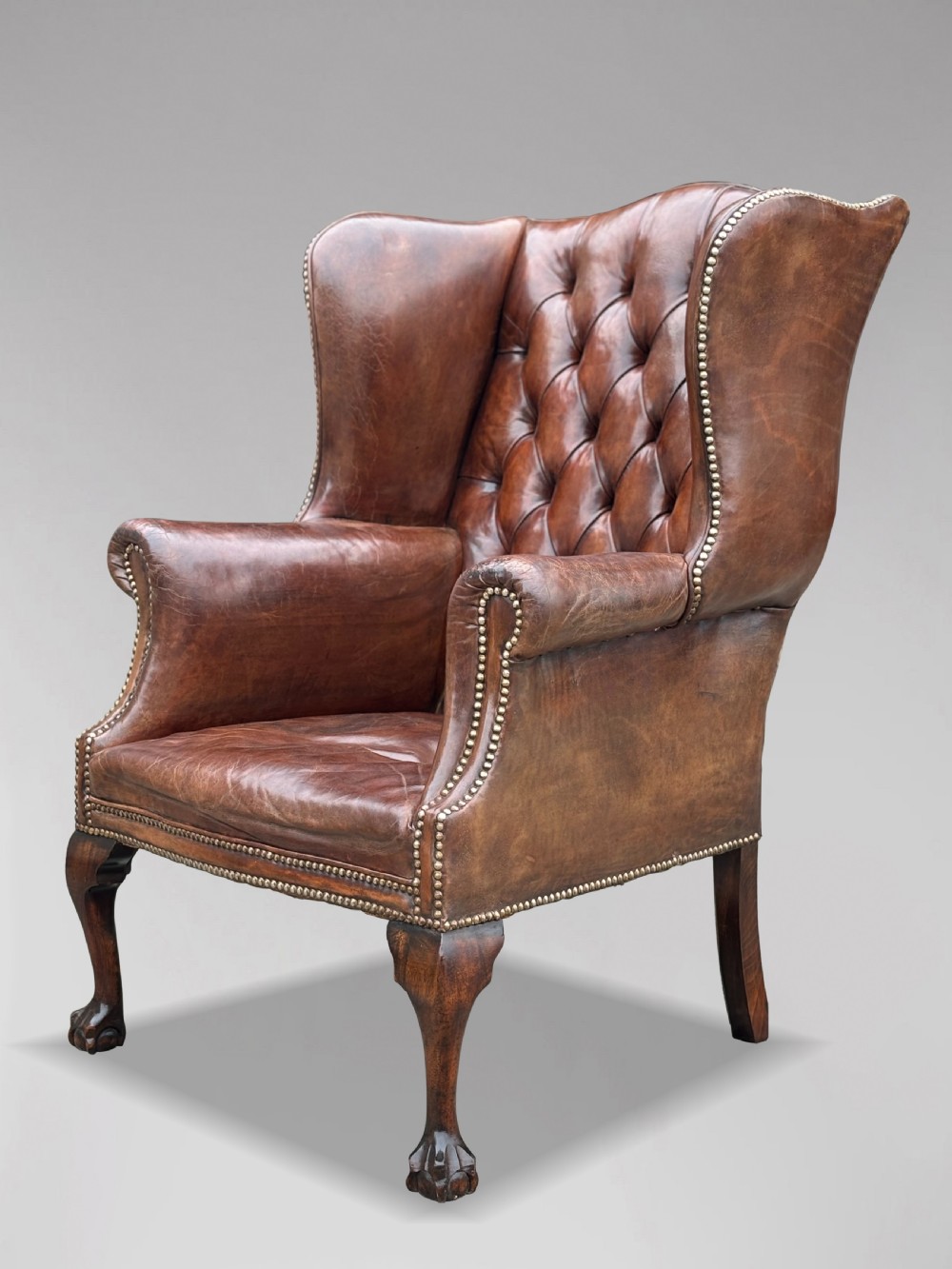 A 19th-century deep brown leather wingback armchair featuring a button-tufted backrest, brass stud trim, and hand-carved mahogany cabriole legs with ball-and-claw feet. View VII