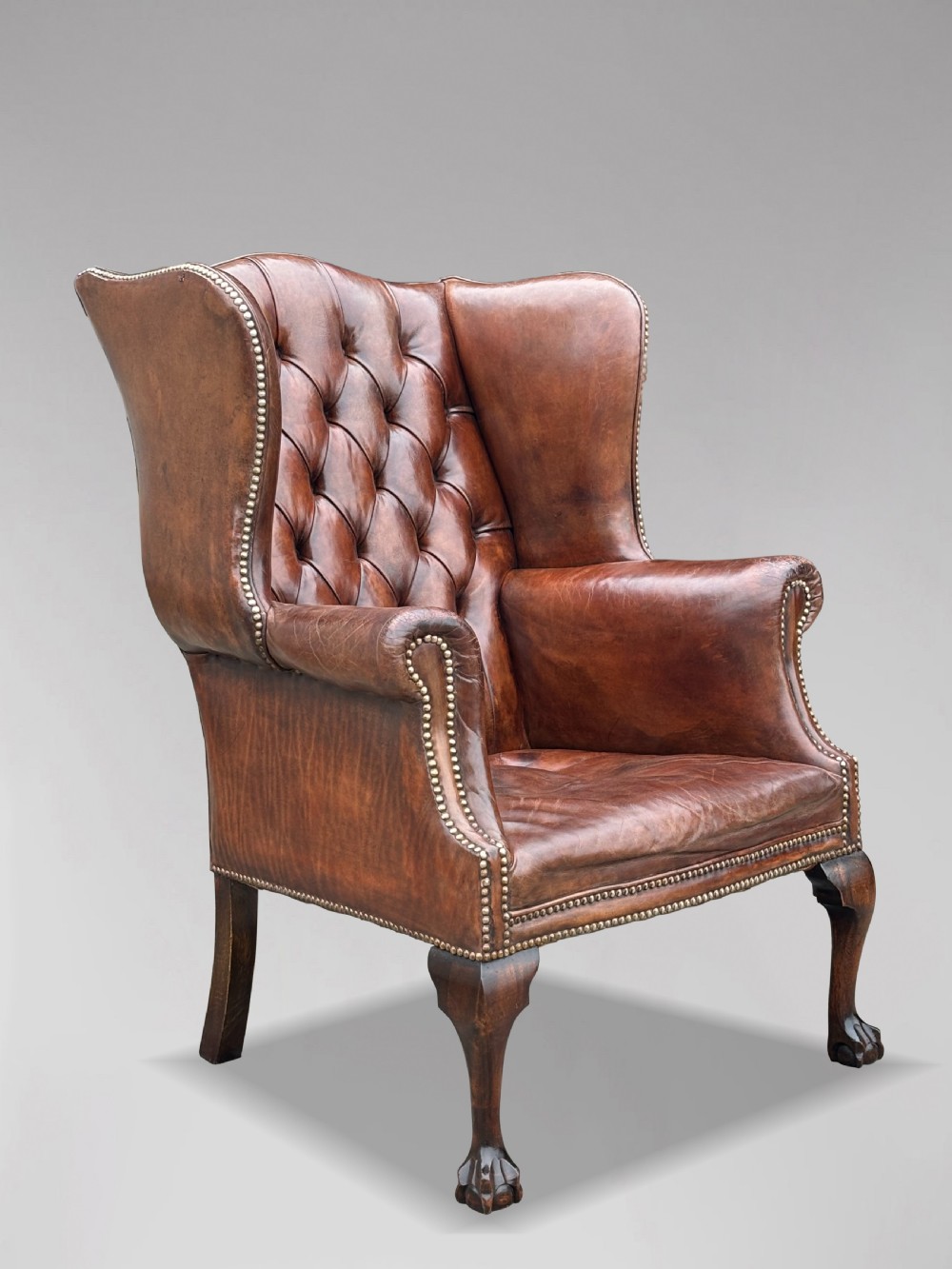 A 19th-century deep brown leather wingback armchair featuring a button-tufted backrest, brass stud trim, and hand-carved mahogany cabriole legs with ball-and-claw feet. View VIII