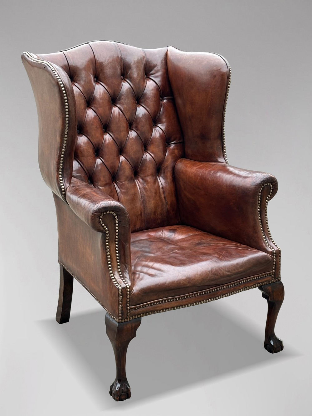 A 19th-century deep brown leather wingback armchair featuring a button-tufted backrest, brass stud trim, and hand-carved mahogany cabriole legs with ball-and-claw feet. View IX