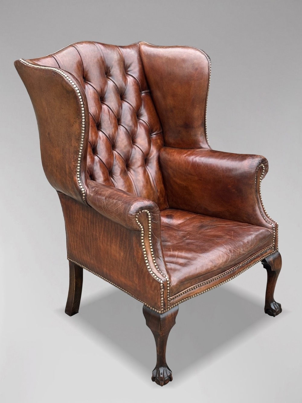 A 19th-century deep brown leather wingback armchair featuring a button-tufted backrest, brass stud trim, and hand-carved mahogany cabriole legs with ball-and-claw feet. View X