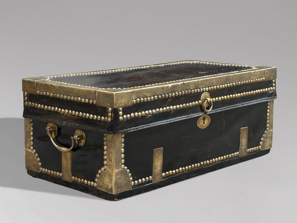 A 19th-century black leather coaching trunk featuring brass corner mounts, brass studding along the edges, side handles, and a central keyhole.