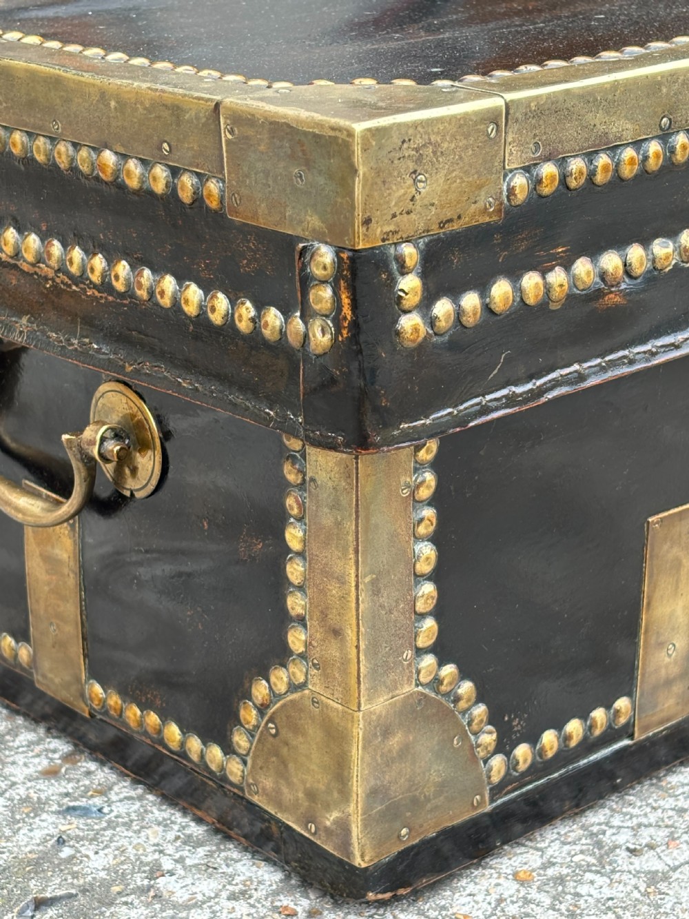 A 19th-century black leather coaching trunk featuring brass corner mounts, brass studding along the edges, side handles, and a central keyhole. View VI