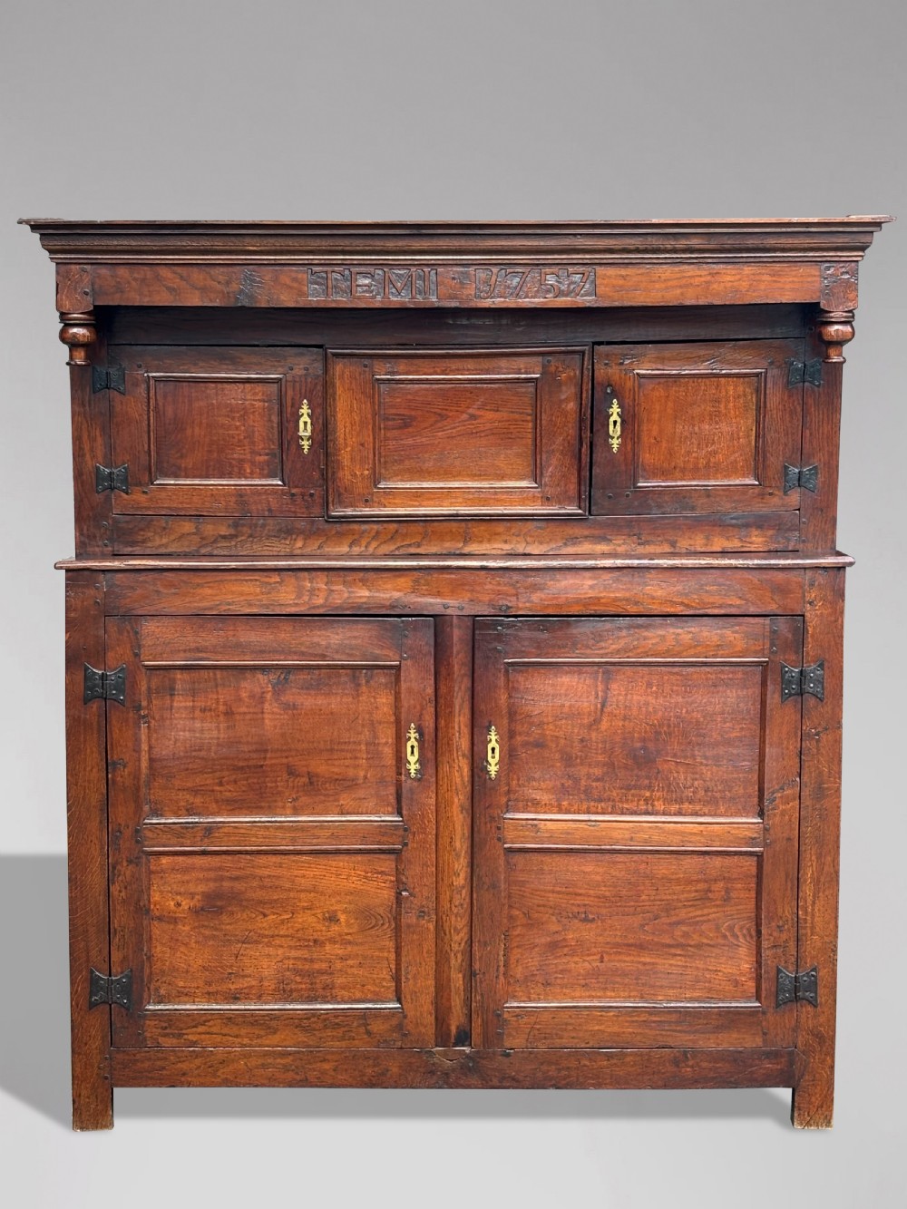 An 18th-century dark elm court cupboard with carved date inscription, panelled doors, brass escutcheons, and iron hinges.