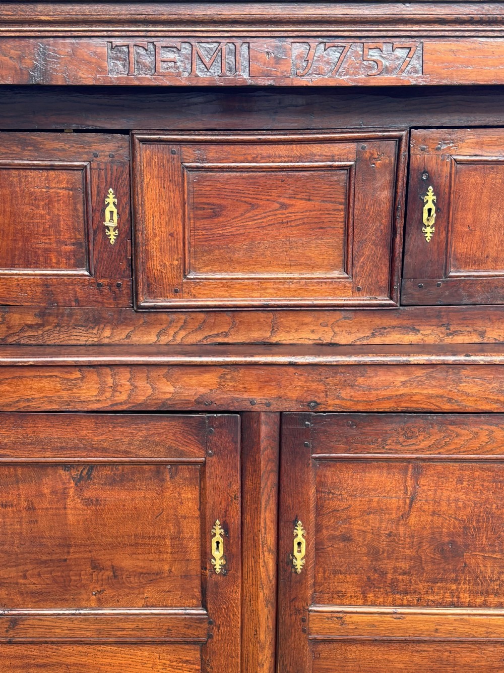 An 18th-century dark elm court cupboard with carved date inscription, panelled doors, brass escutcheons, and iron hinges. View V