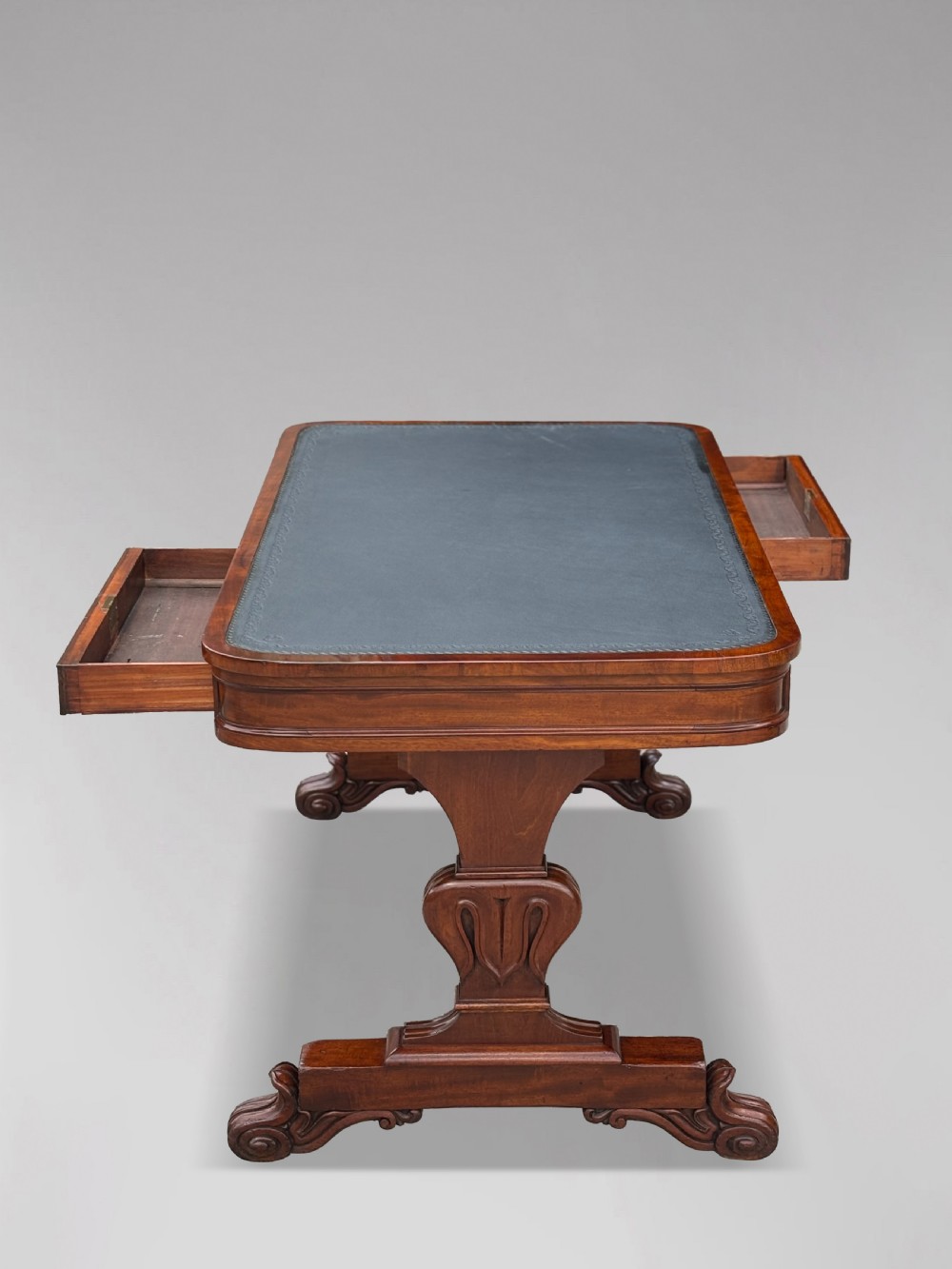 A 19th-century William IV mahogany writing table featuring a dark leather inset top, frieze drawers, carved trestle supports, and scroll feet. View IV