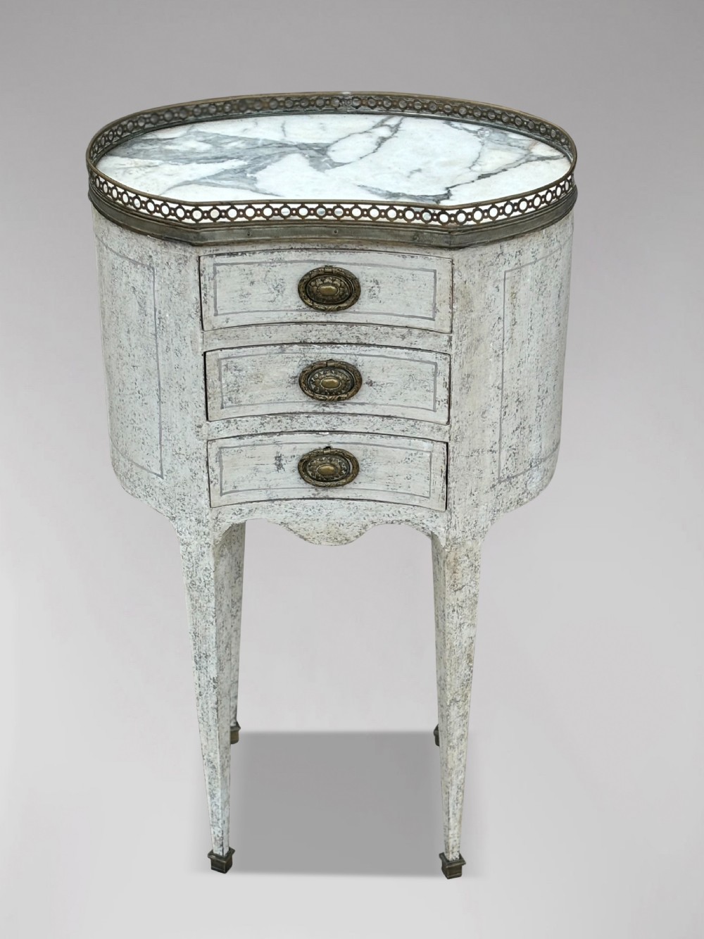 An early 20th-century French Louis XVI style oval night table with distressed grey paintwork, three small drawers, brass pull handles, a marble inset top, and a brass gallery rail. View II