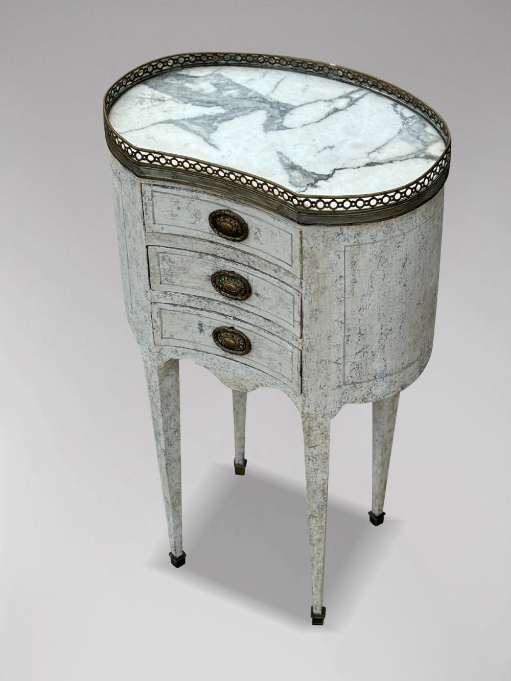 An early 20th-century French Louis XVI style oval night table with distressed grey paintwork, three small drawers, brass pull handles, a marble inset top, and a brass gallery rail. View III