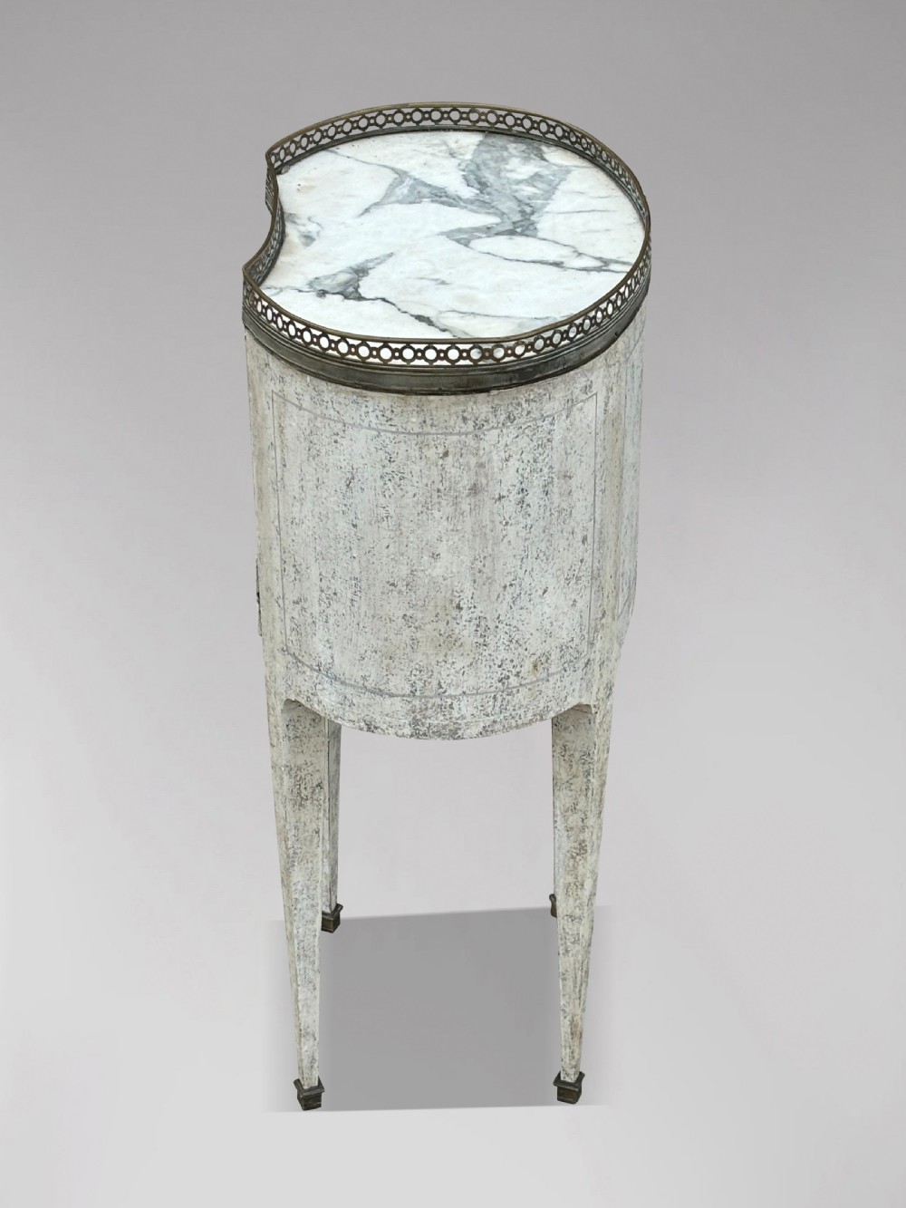 An early 20th-century French Louis XVI style oval night table with distressed grey paintwork, three small drawers, brass pull handles, a marble inset top, and a brass gallery rail. View V