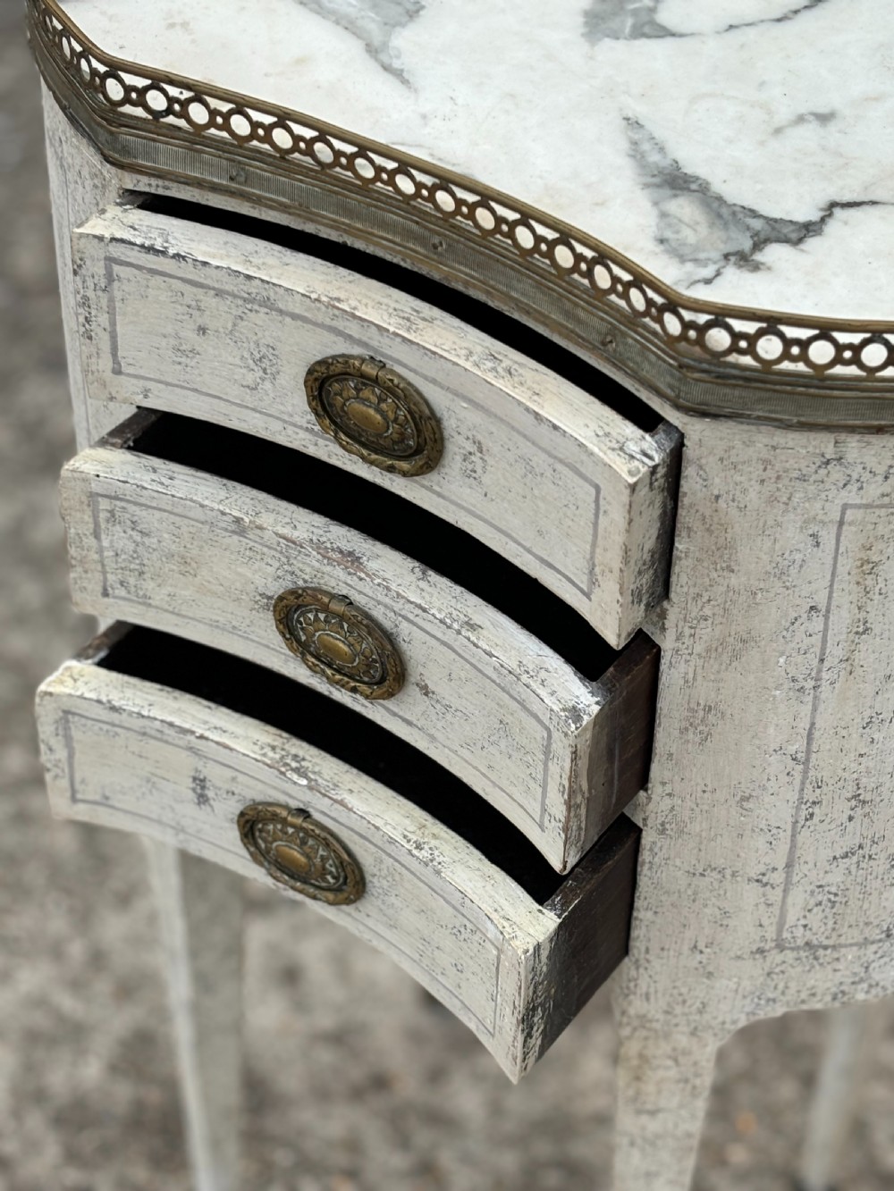 An early 20th-century French Louis XVI style oval night table with distressed grey paintwork, three small drawers, brass pull handles, a marble inset top, and a brass gallery rail. View VI