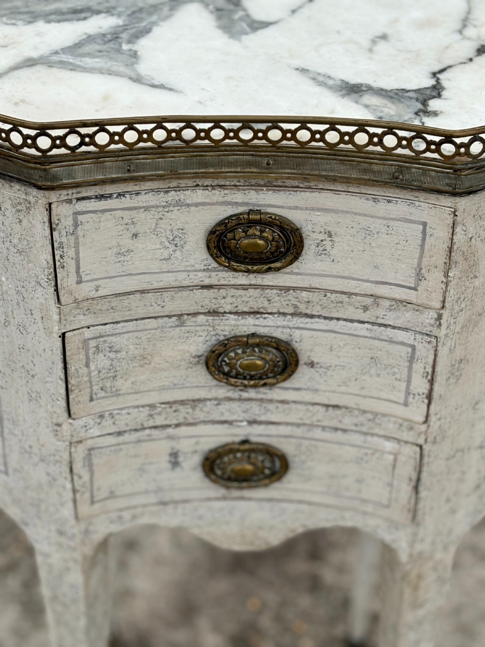 An early 20th-century French Louis XVI style oval night table with distressed grey paintwork, three small drawers, brass pull handles, a marble inset top, and a brass gallery rail. View VII