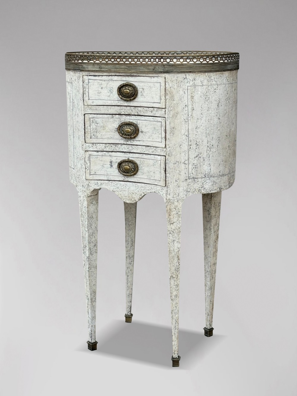An early 20th-century French Louis XVI style oval night table with distressed grey paintwork, three small drawers, brass pull handles, a marble inset top, and a brass gallery rail. View VIII
