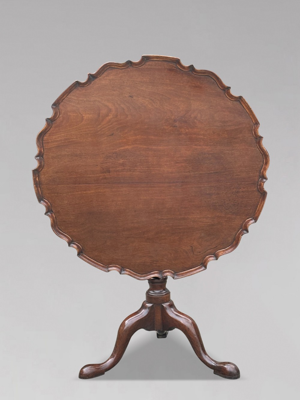 An 18th-century George III mahogany tilt-top tripod table shown in the vertical position with a carved pie-crust rim, turned pedestal, and cabriole legs.