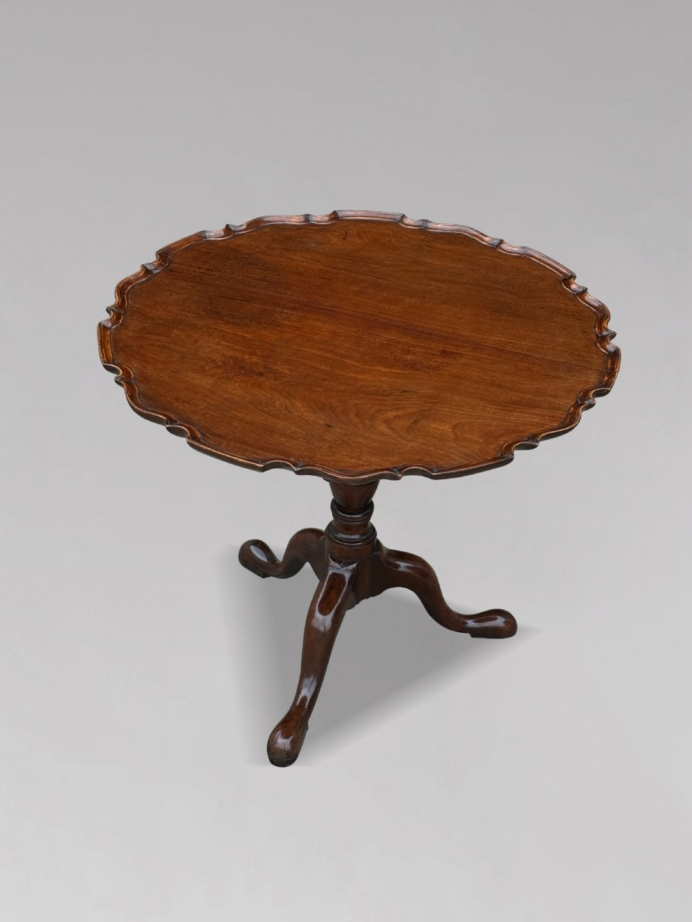 An 18th-century George III mahogany tilt-top tripod table shown in the vertical position with a carved pie-crust rim, turned pedestal, and cabriole legs. View XI