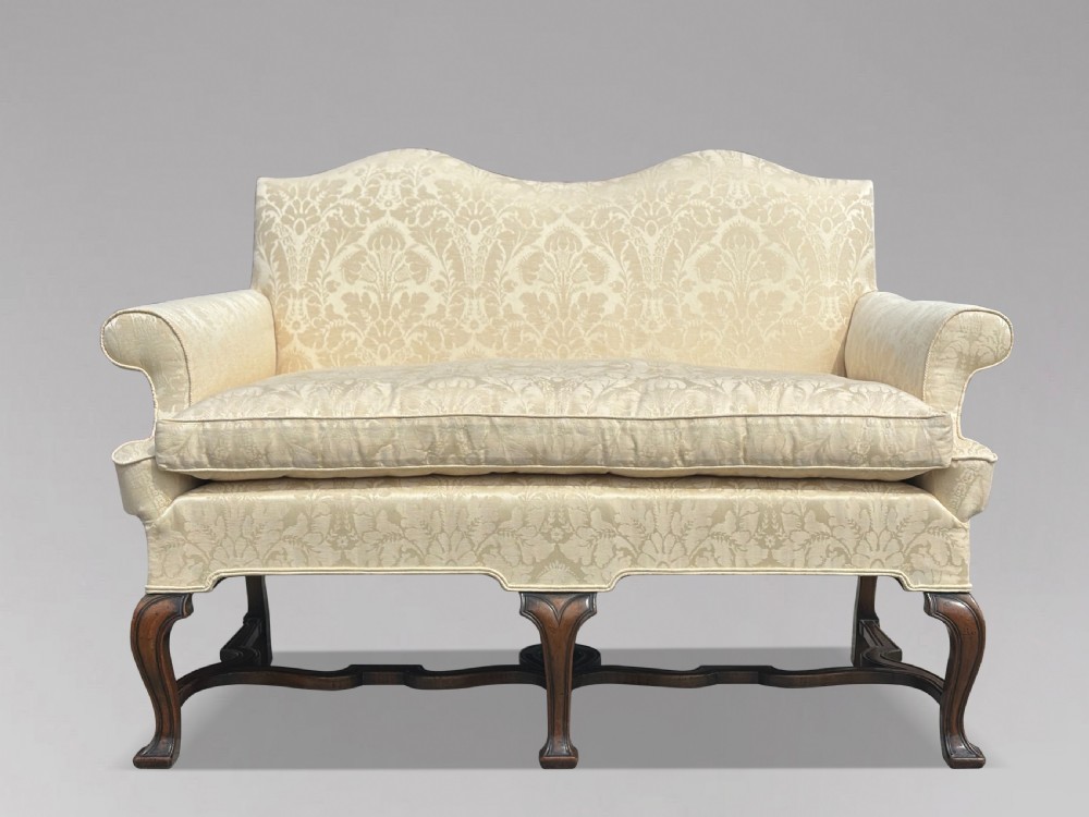 An Edwardian Queen Anne style settee upholstered in cream damask with carved walnut cabriole legs and a double-arched back.