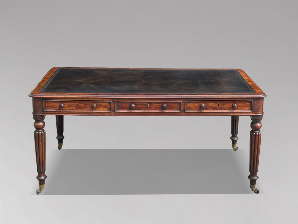 An 18th-century mahogany library table featuring a black leather top insert, frieze drawers with wooden knob handles, and turned, fluted legs on brass castors.