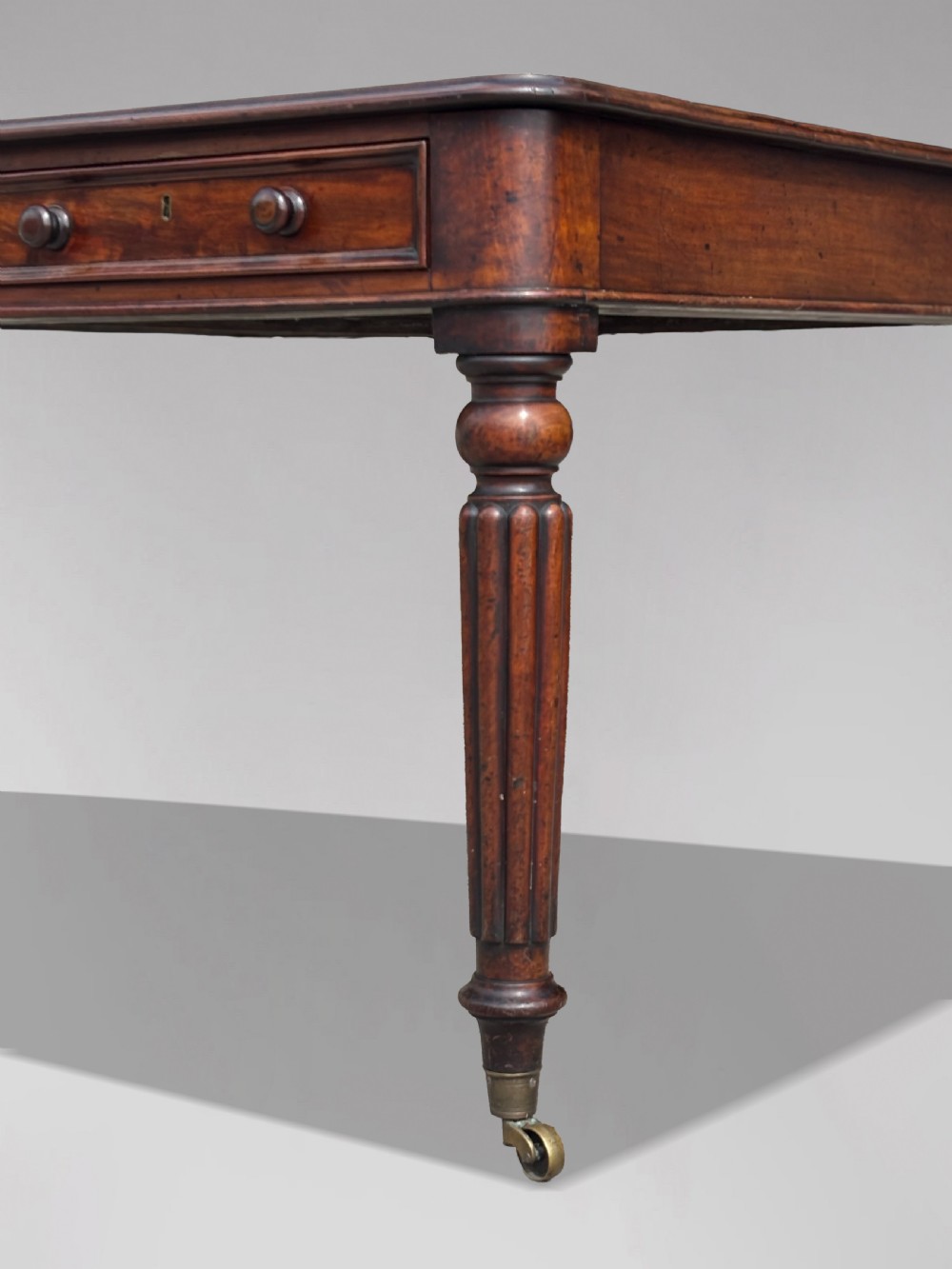 An 18th-century mahogany library table featuring a black leather top insert, frieze drawers with wooden knob handles, and turned, fluted legs on brass castors. View VI
