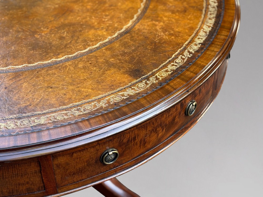 A 19th-century Regency mahogany drum table featuring a circular leather inset top, frieze drawers with brass ring handles, and a carved pedestal with four splayed legs on brass castors. View V